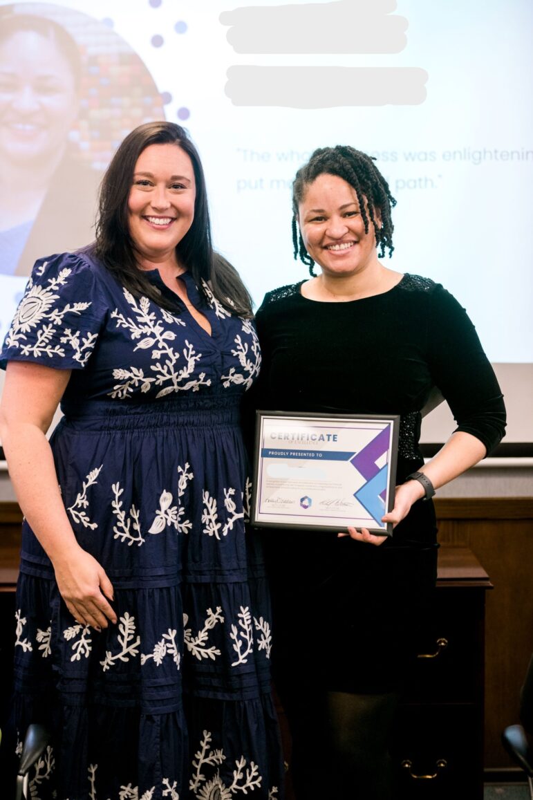 One of our honorees wears a black velvet dress. She's smiling at the camera and holding her certificate of excellence. Standing next to her is financial coach Nikki Whetsell. Nikki is also smiling into the camera and is wear and navy blue dress with white, floral embroidery.