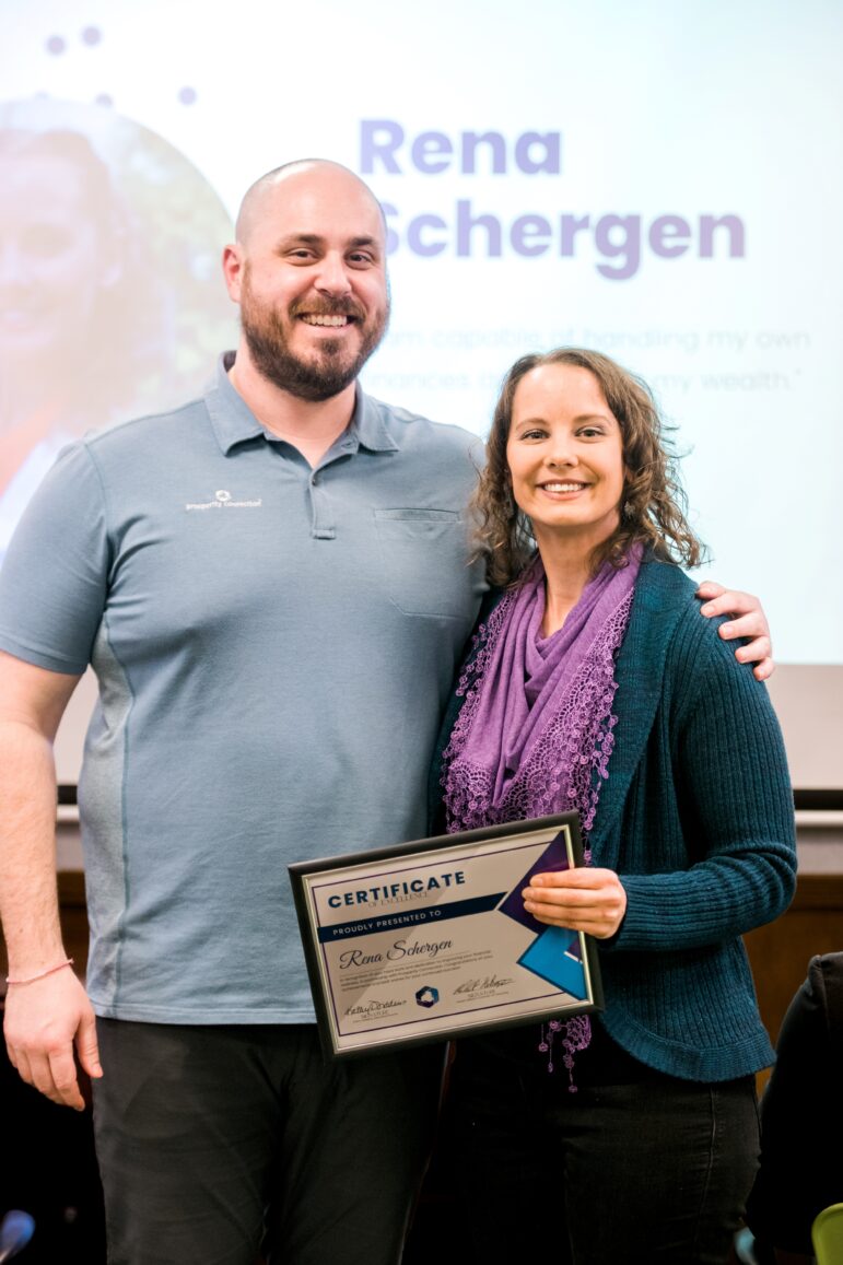 Rena Schergen wears a turquoise cardigan and purple scarf. She's smiling into the camera and holding her certificate of excellence. Standing next to her is financial coach Ben Feimer. He's smiling and wearing a blue-grey polo shirt.