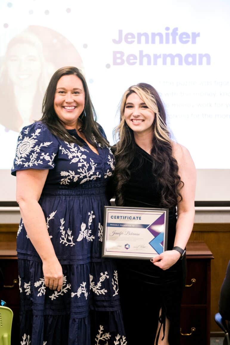 Jennifer Behrman wears a one-shoulder, black velvet dress. She's smiling at the camera and holding her certificate of excellence. Standing next to her is financial coach Nikki Whetsell. Nikki is also smiling into the camera and is wear and navy blue dress with white, floral embroidery.