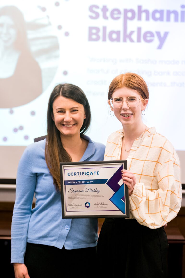 Stephanie Blakley is wearing a cream button-up blouse with an orange square pattern, and a black skirt. She has square glasses with clear rims and wears braces. She is smiling at the camera and holding her certificate of excellence. Standing next to her is financial coach Sasha Moore. Sasha also smiles at the camera and is wearing a light blue cardigan with a black skirt.