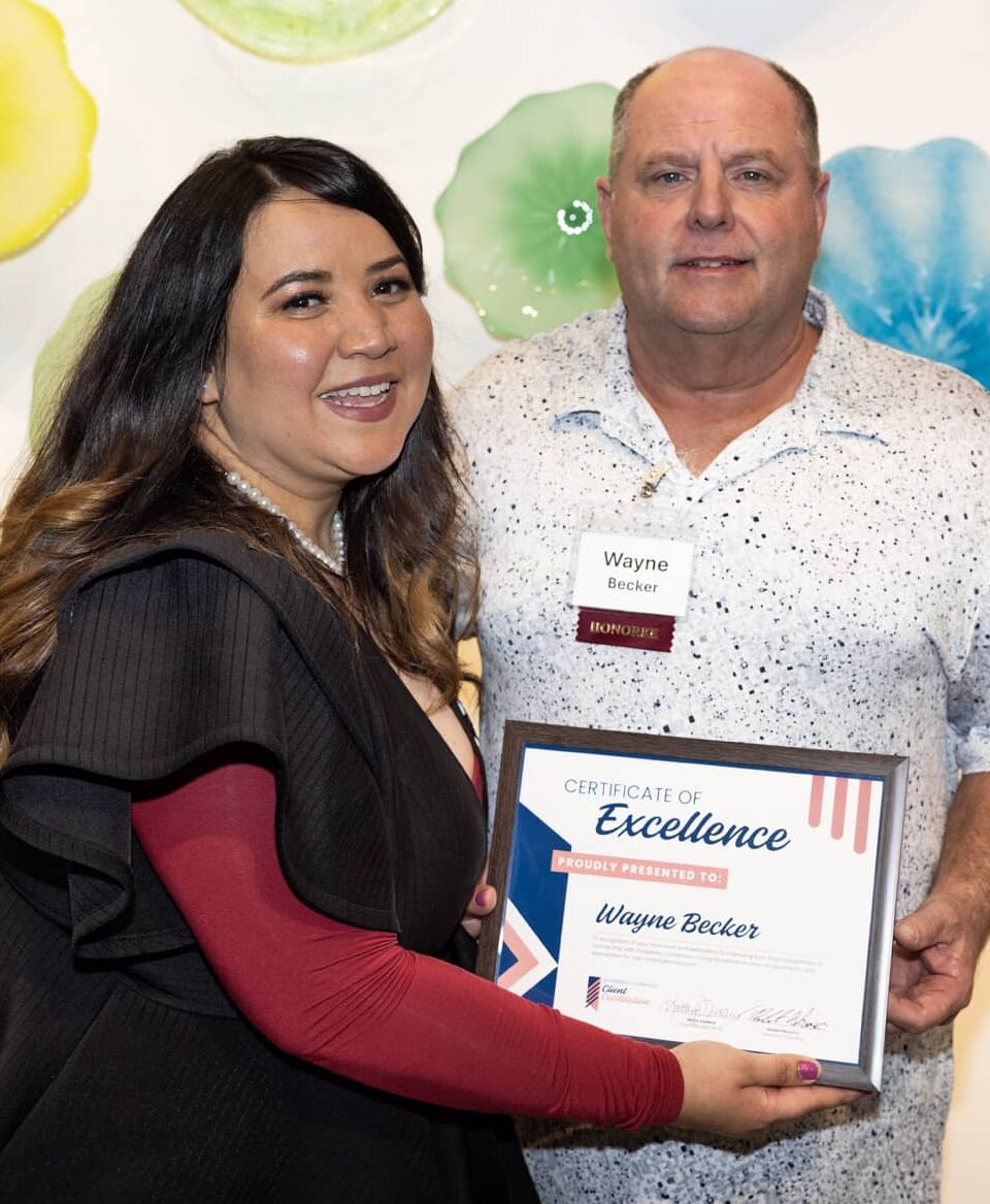 Wayne Becker posing for a photo while standing next to his financial coach, Veronica Guerrero. They are holding his Certificate of Excellence and smiling.
