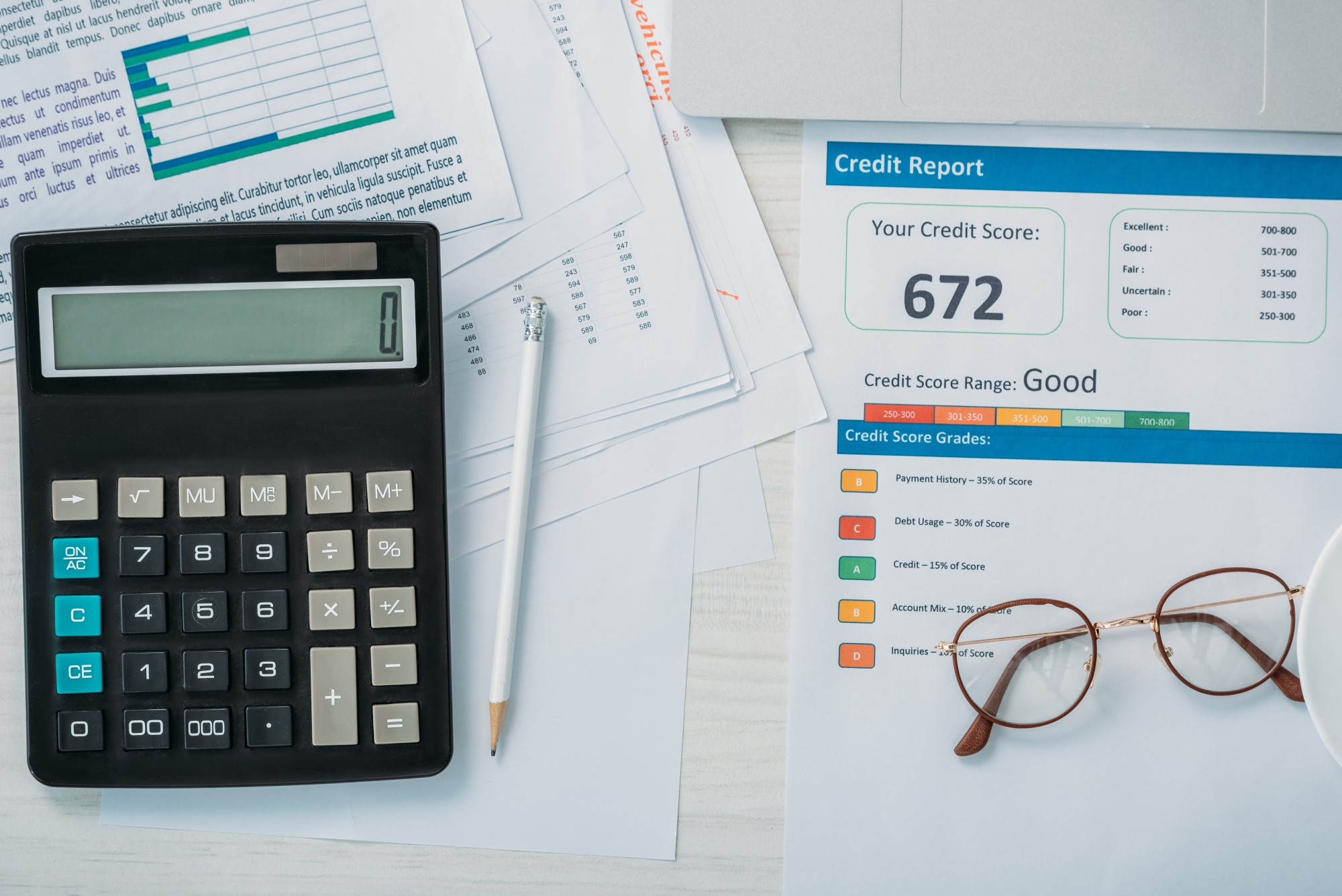 A credit report is laid on a table with a pair of glasses resting on top. To the left of the credit report is a pile of financial documents, a calculator, and a pencil.