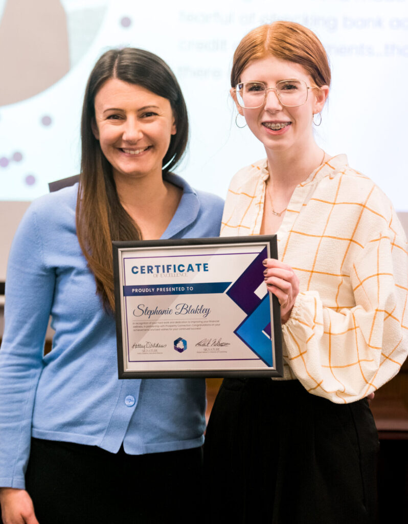 Stephanie Blakley is wearing a cream button-up blouse with an orange square pattern, and a black skirt. She has square glasses with clear rims and wears braces. She is smiling at the camera and holding her certificate of excellence. Standing next to her is financial coach Sasha Moore. Sasha also smiles at the camera and is wearing a light blue cardigan with a black skirt.