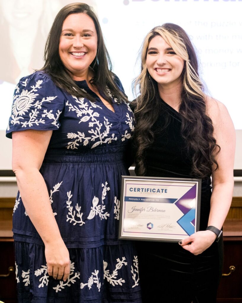 Jennifer Behrman wears a one-shoulder, black velvet dress. She's smiling at the camera and holding her certificate of excellence. Standing next to her is financial coach Nikki Whetsell. Nikki is also smiling into the camera and is wear and navy blue dress with white, floral embroidery.