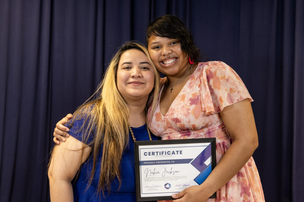 Nakia Jackson stands with her financial coach Veronica Guerrero as they both smile warmly and proudly hold up Nakia's Certificate of Excellence.