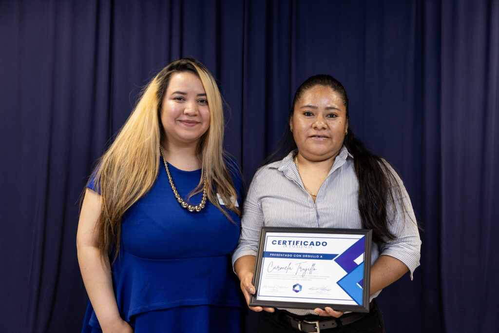 Carmella Tiburcio stands with her financial coach Veronica Guerrero as they both smile warmly and proudly hold up Carmela's Certificate of Excellence.