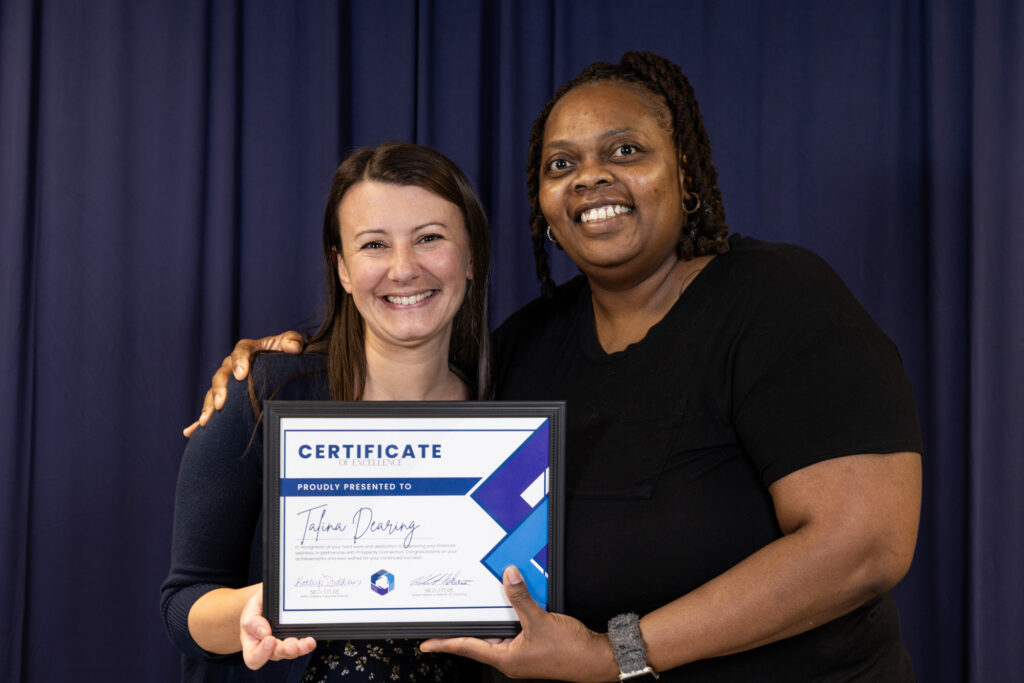 Talina Dearing stands with her financial coach Sasha Moore as they both smile warmly and proudly hold up Talina's Certificate of Excellence.