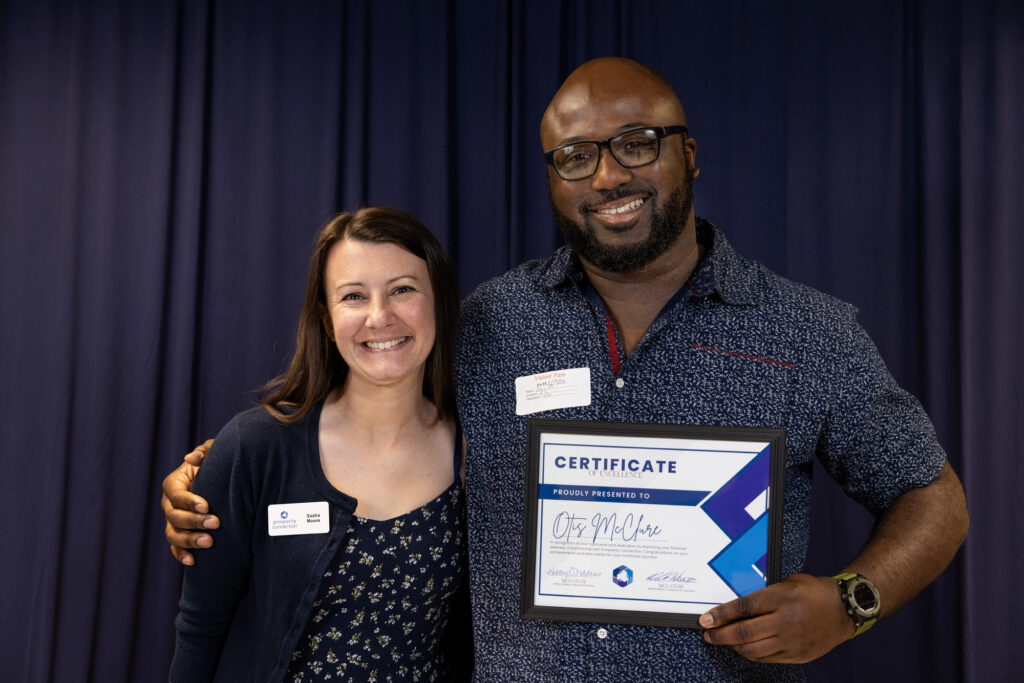 Otis McClure stands with his financial coach Sasha Moore as they both smile warmly and proudly hold up Otis's Certificate of Excellence.